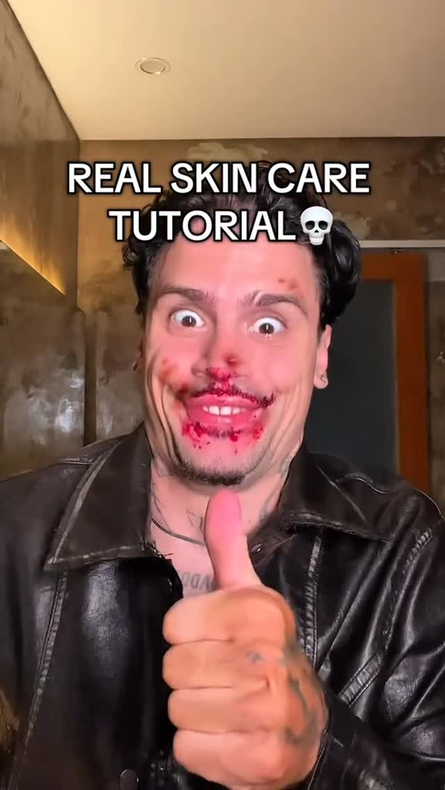The image is a selfie-style shot of a man with short hair and visible tattoos on his arm. He's wearing a black leather jacket. His face is smeared with what appears to be red liquid, covering his mouth and nose, and he's giving a thumbs up with a wide, excited expression. Above him, text reads: 'REAL SKIN CARE TUTORIAL ☠️'.