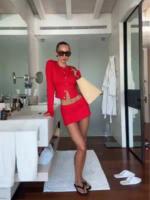 A woman in a red outfit, including a cardigan and mini skirt, stands in a bathroom setting with a mirror and sink in the background.
