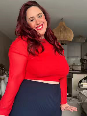 A medium shot of a woman with red hair, wearing a red top and a blue skirt. She is smiling, and the background is a living room.