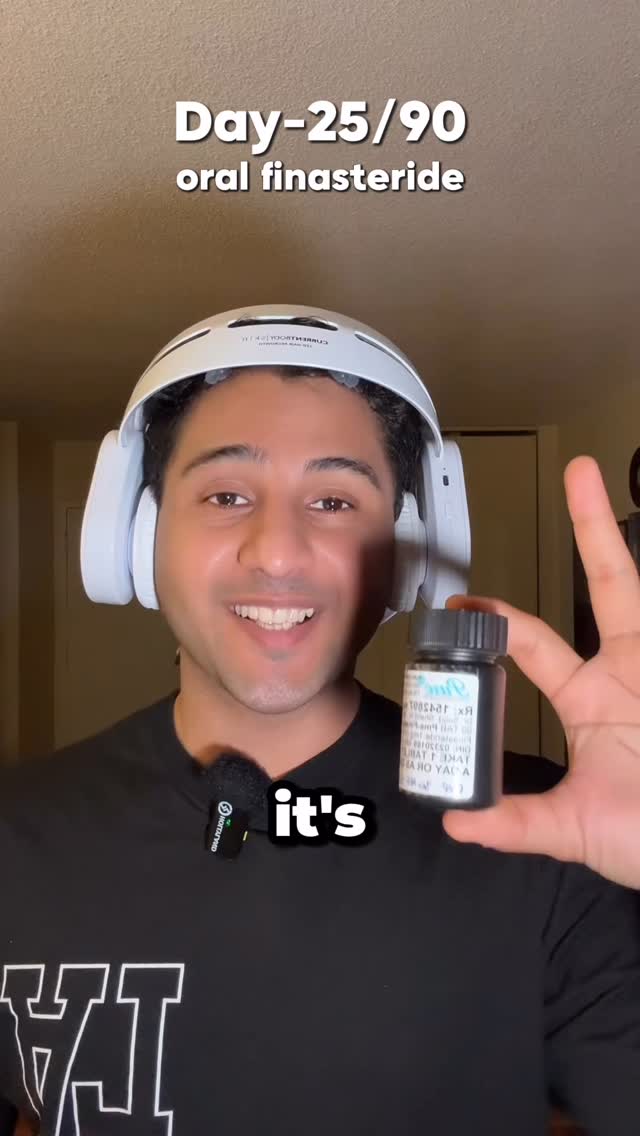 A young man with dark hair and light skin is smiling at the camera. He is wearing a black t-shirt and a white headset. The background is a plain light-colored wall with a door partially visible on the right. Above the man's head, the text states, "Day-25/90 oral finasteride". In his right hand, he holds a small bottle of medication over his chest, with some printed information visible on the label. A black label is at the bottom of the image that states "it's". A microphone is clipped to his shirt.