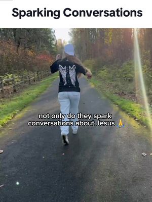 A person with long hair, wearing a white cap and shirt with a graphic on the back depicting wings, is running down a paved path through a lush green area. The text overlay reads "Sparking Conversations" at the top and "not only do they spark conversations about Jesus" near the bottom. The background is blurred, featuring trees and sunlight.