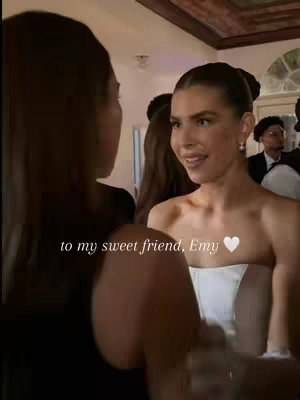 The cover image shows two women in an indoor setting, likely at an event. One woman is in focus, and the other is partially visible, with their back facing the camera. The woman in focus has a warm, friendly expression and is wearing what appears to be a gown or formal attire. Text overlay appears at the bottom with text about Emy.