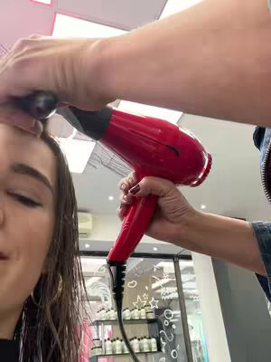 A woman is having her hair styled with a red hairdryer in a salon. The image is a close-up, with the woman's face on the left side of the frame and the stylist's hands holding the hairdryer on the right. The background is blurred, featuring a glimpse of a salon interior with lights and products.