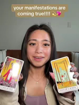 A young woman with long dark hair holds up two tarot cards while looking at the viewer and smiling. A text box across the top says "your manifestations are coming true!!!! 💫💖". The background is a blurred room setting.