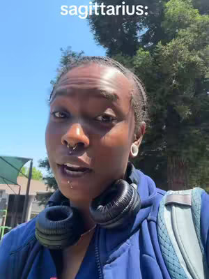 A close-up shot of a person with dark skin, wearing a blue zip-up jacket and headphones around their neck. The person is looking at the camera and talking with a serious expression. The background shows a sunny day with trees and some buildings.