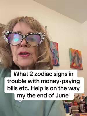 A woman with blonde hair and decorative glasses is centered in the image, looking directly at the camera. Text overlay in a white box appears at bottom of the shot. Background art is hanging behind the woman.