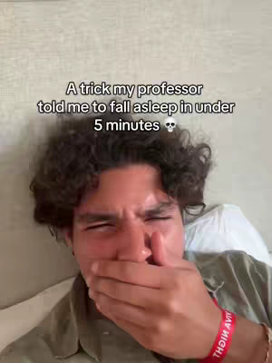 A selfie of a young man in a bed, with text overlayed. He is covering his mouth with his hand.. The background shows a light-colored blanket and pillow, which provides a soft backdrop. The man's expression indicates surprise or slight amusement.