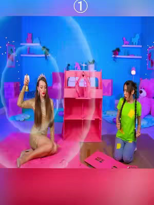 Two young women are positioned in a brightly lit room with a whimsical aesthetic. One, dressed in a gold dress and tiara, appears to be using a prop, while the other, in a green top and blue jeans, looks on in surprise. A large, pink dollhouse structure is in the background, and the color palette is dominated by shades of pink, blue, and purple. Decorative items are arranged on shelves, contributing to a playful and imaginative vibe.