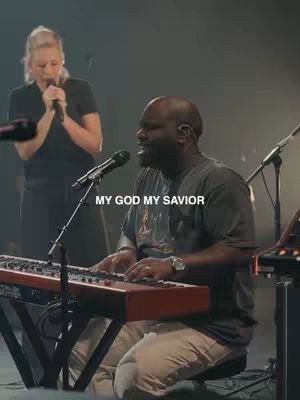 A live music scene with a man playing a keyboard and singing, another person with a microphone is behind him. The room is dimly lit.