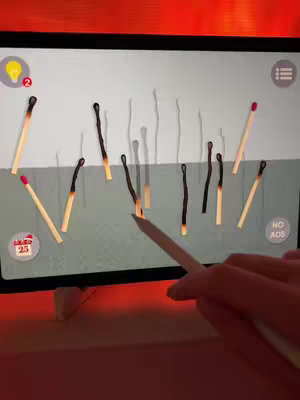 A tablet screen displays a game interface with several matchsticks arranged in a seemingly random manner against a light gray background. The matches vary in orientation, some are vertical and some angled, with some appearing to be burnt at the tip. A hand holding a white stylus pen indicates the player's interaction with the game. The scene is illuminated by a warm orange glow.