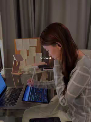A person is seated at a glass desk, working on a laptop and a tablet. The scene is illuminated by a soft light, and the person is wearing a light colored sweater. There is a word in the center of the image in a small font.