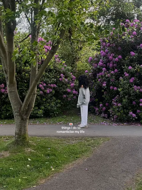 A person stands with their back to the camera, in a lush outdoor setting. They are wearing a gray sweatshirt and white pants. The background is filled with green foliage, flowering bushes with purple blooms, and a paved pathway. A small, rectangular graphic at the bottom contains text.