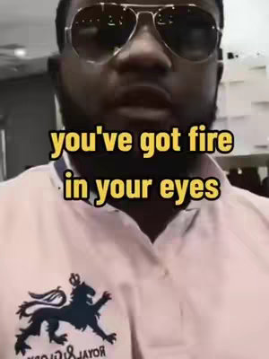 A medium close-up shot featuring a man with dark skin wearing aviator sunglasses. The man is looking up slightly towards the camera. He is wearing a light-colored shirt with a dark emblem on the chest. Overlaid, in large yellow font, is the text 'you've got fire in your eyes' and in smaller yellow letters under that, 'in your eyes'.