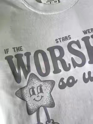 A close-up shot of a white t-shirt with a graphic print. The design features the words "IF THE STARS WORSHIP" in large, gray, outlined letters. Below, a cartoon star with a smiling face and stick legs is present. The composition focuses on the details and the message of the shirt.