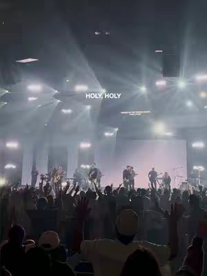 A live worship service with a band playing on stage. Silhouetted figures of people in the audience with their hands raised. Stage lights create a soft, slightly overexposed effect.