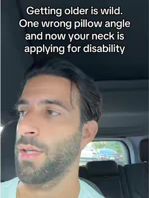 A man is filmed from the torso up while sitting inside a vehicle, likely a car. The man is light-skinned, has dark hair, and a full beard. The text overlay on the image reads: "Getting older is wild. One wrong pillow angle and now your neck is applying for disability".The background is blurred, with other cars and greenery visible through the windows.