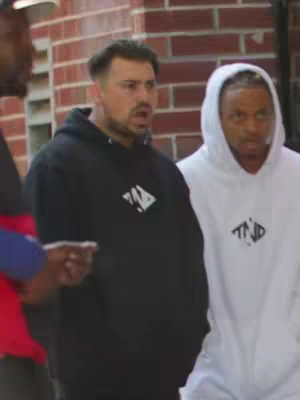 The image captures three men in an outdoor setting against a brick wall. The main focus is on the man in the center, who is wearing a black hoodie. Another man to the right is wearing a white hoodie. Both hoodies feature a logo. The man on the left is partially visible. The overall composition suggests a candid moment or scene.