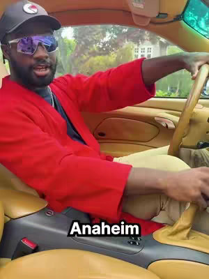 A man in a red jacket and sunglasses is driving a car. The car’s interior is tan and yellow, and the man is wearing a black cap. Text that says 'Anaheim' is overlaid at the bottom of the image.