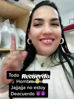 The image is a selfie of a woman smiling and pointing upwards with her right index finger. Her dark hair is pulled back from her face, and she wears large hoop earrings and a white collared top. There is a text overlay on the bottom portion of the image with the colors black and pink. The background includes a partial view of shelves.