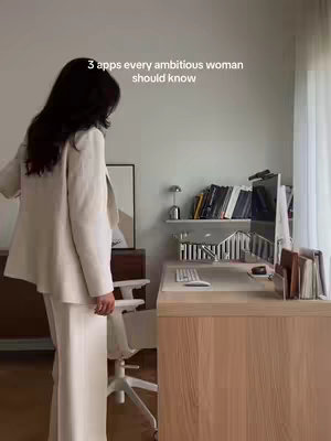 A woman in a cream-colored suit stands in an office setting, her back to the camera. In the background, there are office supplies like a computer setup and some books. Overlaid text at the top suggests the content discusses apps for ambitious women. The overall tone is clean and organized.