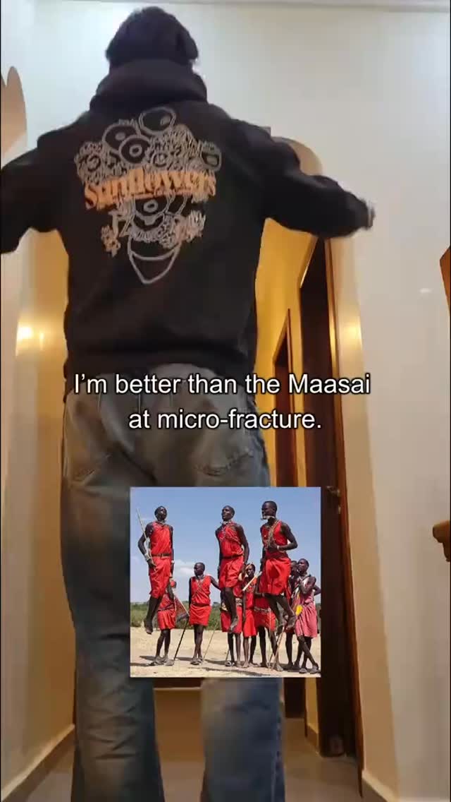 The cover image features a person from the back, standing in the center of the frame with their back to the camera. They are wearing a black hooded sweatshirt with a detailed design on the back. The bottom half of the person is visible, showing them wearing dark jeans. In the lower portion of the frame, a smaller image displays a group of Maasai people, with red traditional clothing, in a field of a bright blue sky. The background consists of a hallway with a yellow wall and brown door frame, with a text overlay that reads, 'I'm better than the Maasai at micro-fracture.’