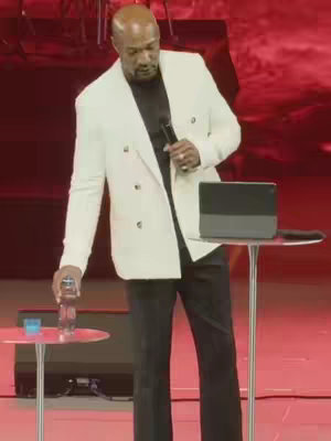 The cover image shows a man in a white blazer standing on a stage. He holds a microphone in one hand and has a bottle of water and a glass of water on a small table next to him. The background is red with a subtle gradient. The lighting suggests a stage setting, and the composition is centered and focused on the man.