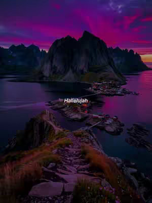 A scenic landscape photo featuring a body of water, a series of dark, imposing mountains, and a pathway in the foreground leading towards a small settlement lit up at the water's edge. The sky transitions from shades of deep purple to orange and yellow, suggesting either a sunset or sunrise. The mountains are dark and silhouetted against the vibrant sky.