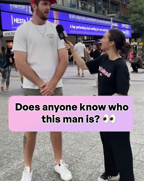 A woman in a black t-shirt holding a microphone interviews a man standing in front of a city backdrop. A pink bubble is overlaid on the image with text: "Does anyone know who this man is?" with eye emojis.