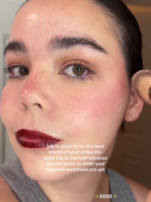 A close-up shot features a fair-skinned person's face, heavily made up with dark lipstick and prominent blush. The person, possibly a woman, has dark hair pulled back, revealing high cheekbones and expressive eyes. The focus is on the face, with a makeup brush touching one cheek. Overlaid on the image is text with motivational language, adding to the image's message.