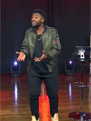 A man in a green bomber jacket and black outfit is on stage, gesturing broadly while speaking. The background is a dark stage setting with red and purple lighting.