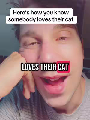 A close-up shot of a man with fair skin, smiling and looking at something off-camera. The background is a blurry interior. Text overlays read 'Here's how you know somebody loves their cat' and 'LOVES THEIR CAT'. The man is wearing a gray shirt.