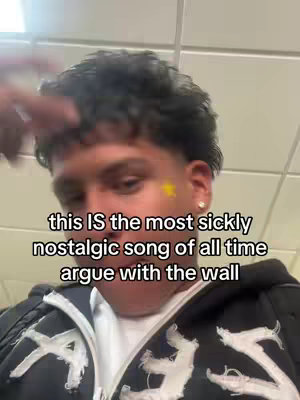 A selfie-style shot featuring a young man from the chest up. He has short, dark, curly hair and is wearing a black zip-up hoodie with white detailing. A yellow star sticker is adhered below his left ear. The background appears to be a plain white tiled ceiling.