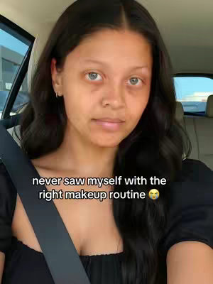 A woman with long, dark hair sits in a car. The text 'never saw myself with the right makeup routine' is overlaid on the image. The woman is gazing forward with a neutral expression, looking at the camera. She is seated in the driver's seat of a vehicle.