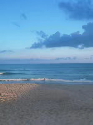 A tranquil beach scene at dusk or dawn, featuring a sandy shore in the foreground that transitions into the calm, rippling ocean. The sky is a gradient of blues, with darker clouds adding texture and depth. The composition is balanced, with soft lighting suggesting a serene atmosphere.