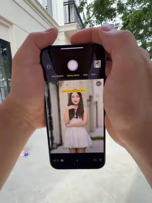 A person is holding an iPhone 15 Pro Max, with the camera app open, showing a woman in a white dress. The phone and hands are the primary focus.
