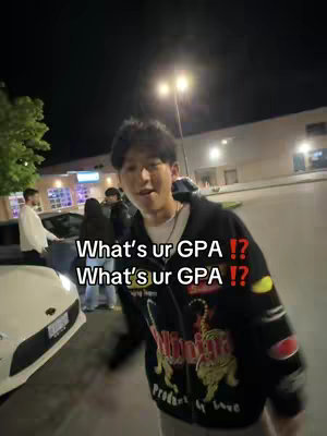An outdoor shot at night. The main subject, a young man with dark hair, is in the center, facing the camera. He wears a jacket and is slightly smiling at the camera. Two lines of white text are overlayed on the image: "What's ur GPA !?" The background includes bright streetlights and partially visible buildings and some people.