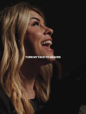 A woman with shoulder-length blonde hair looks upwards with a slight smile and her mouth open as if singing. Text overlayed reads 'TURN MY FACE TO HEAVEN'. The background is dark and out of focus, creating a contrast with the bright lighting on the woman's face.