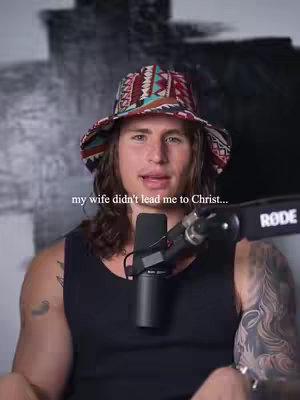 A man with long hair wearing a patterned hat and a black tank top is speaking. A microphone is positioned in front of him. Text overlaid on the image reads 'my wife didn't lead me to Christ...'