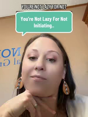 A woman with brown hair and dark makeup is featured in a selfie-style shot, with the camera focused on her face. She has her hand resting on her chin, and her expression appears contemplative. A text overlay in a speech bubble reads, "You're Not Lazy For Not Initiating." The background is a light orange, providing a soft contrast to the woman's features and the text overlay.