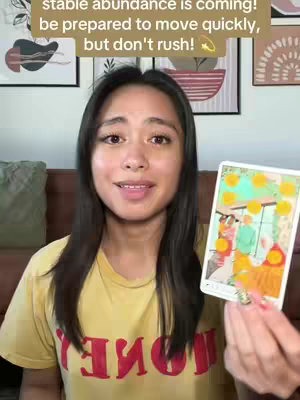 A woman with dark hair and a yellow top holds a tarot card, facing the camera with a slight smile. Behind her are framed artworks, suggesting a home setting. The card shows a colorful illustration with people and golden coins. The overlay text provides a message about abundance.