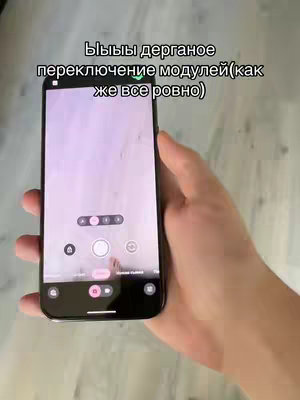 A hand holding a smartphone that is displaying the camera app interface. The phone screen is mostly filled with the camera's preview, showing a portion of the room. Text overlay is added to the top of the screen.