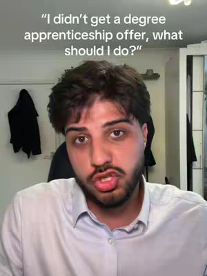 A man with dark hair and a beard looks directly at the camera, his mouth slightly open as if speaking. Above his head appears text in a stylized font. The background is a cluttered room with a white wall and a dark jacket hanging from the wall.