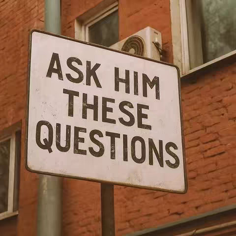 A square, weathered sign reads "ASK HIM THESE QUESTIONS" in bold, dark, capital letters. The sign is white with visible wear and is mounted on a metal post. The background features a red brick wall, a portion of a window, and an air conditioning unit.