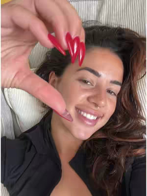 A selfie-style cover image showing a woman lying on a bed with a light-colored, textured headboard. She's smiling, looking directly at the camera. Her right hand is raised, with fingers forming a heart shape. The nails are long and painted red. The lighting is natural, and the background is soft-focused.