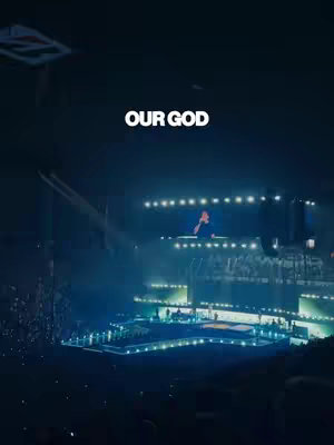 The image shows a concert stage with bright spotlights. The words “OUR GOD” are prominently displayed in white text. The stage is multi-tiered, with visible people in the foreground, and a person speaking in the center.