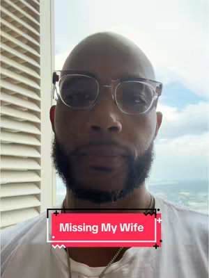 A close-up selfie of a man wearing glasses, with a beard and a white shirt, set against a blurred background of a window and sky. A red and white bar overlaid on the bottom of the image reads, "Missing My Wife."