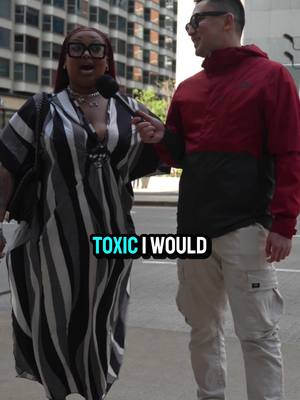 An image from a street interview features two individuals, a woman and a man. The woman is on the left, wearing a striped dress, glasses, and holding a microphone. The man is on the right, wearing a red and black jacket and glasses. In the foreground, overlaid text reads 'TOXIC I WOULD'. The background shows a city street with buildings.