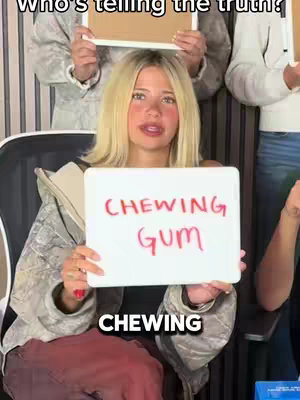 A blonde woman sits in a chair holding a whiteboard that says 'CHEWING GUM'. The text 'CHEWING' appears below it, and two other people hold boards above her, though their text is illegible. The background is blurred.