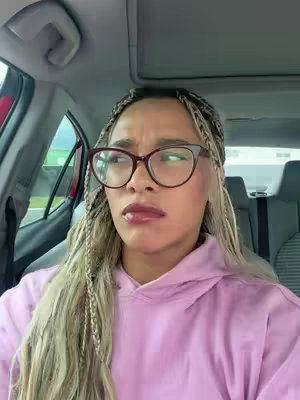 A woman with blonde braids wearing glasses and a pink hoodie is sitting in a car. Her expression is quizzical, and the interior of the car is visible in the background. The lighting is natural.