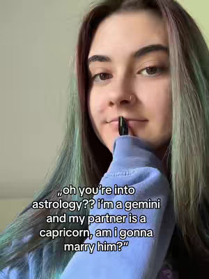 A young woman is centered in the frame, gazing slightly to the side with a neutral expression. Her long hair has streaks of green and reddish-purple, framing her face. She wears a light blue sweatshirt and has a nose ring. She's holding a dark lipstick or eyeliner pencil near her lips. Text overlays are present, offering a question about astrology.